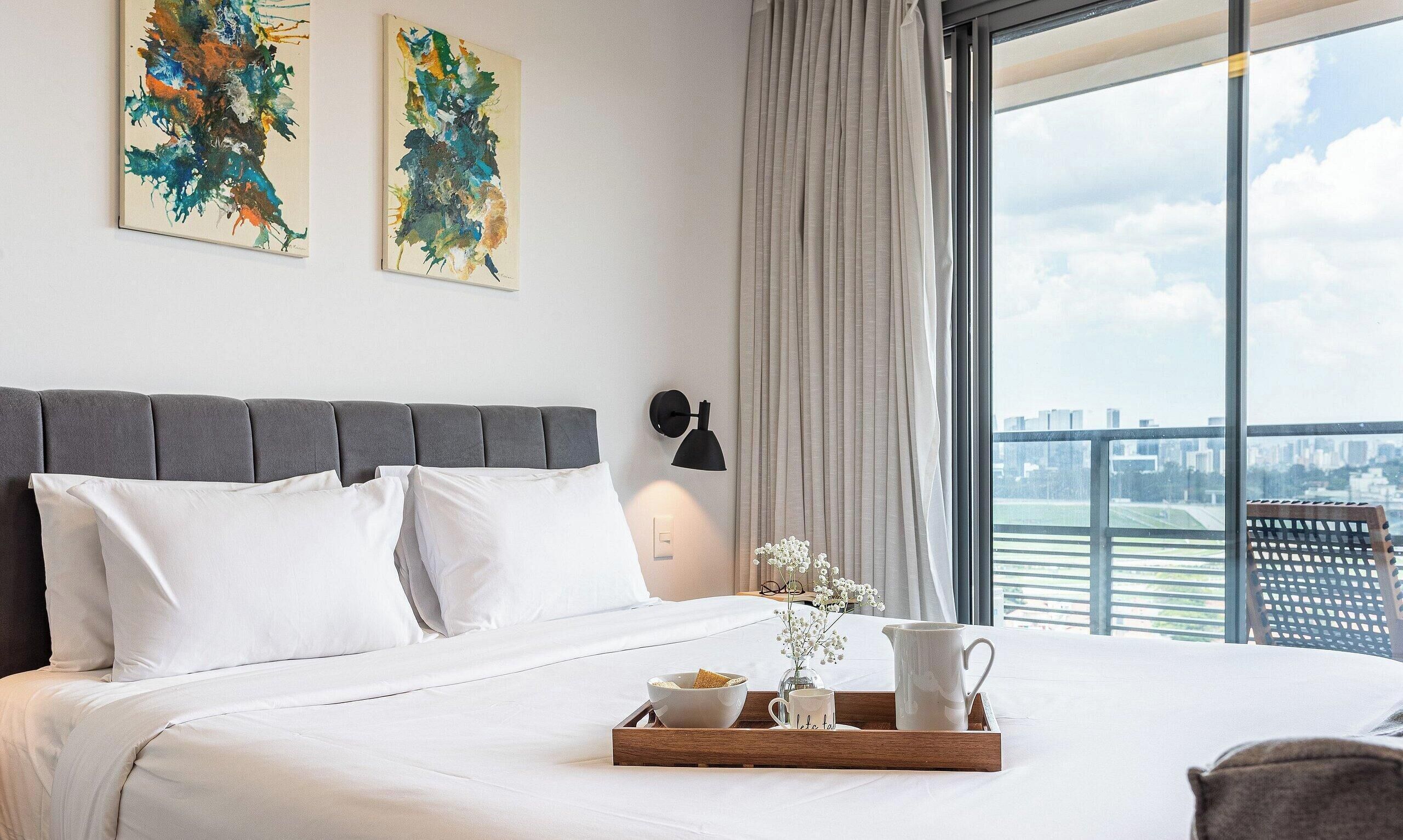A hotel room with a bed, a nightstand, a window with a city view, and artwork on the wall.
