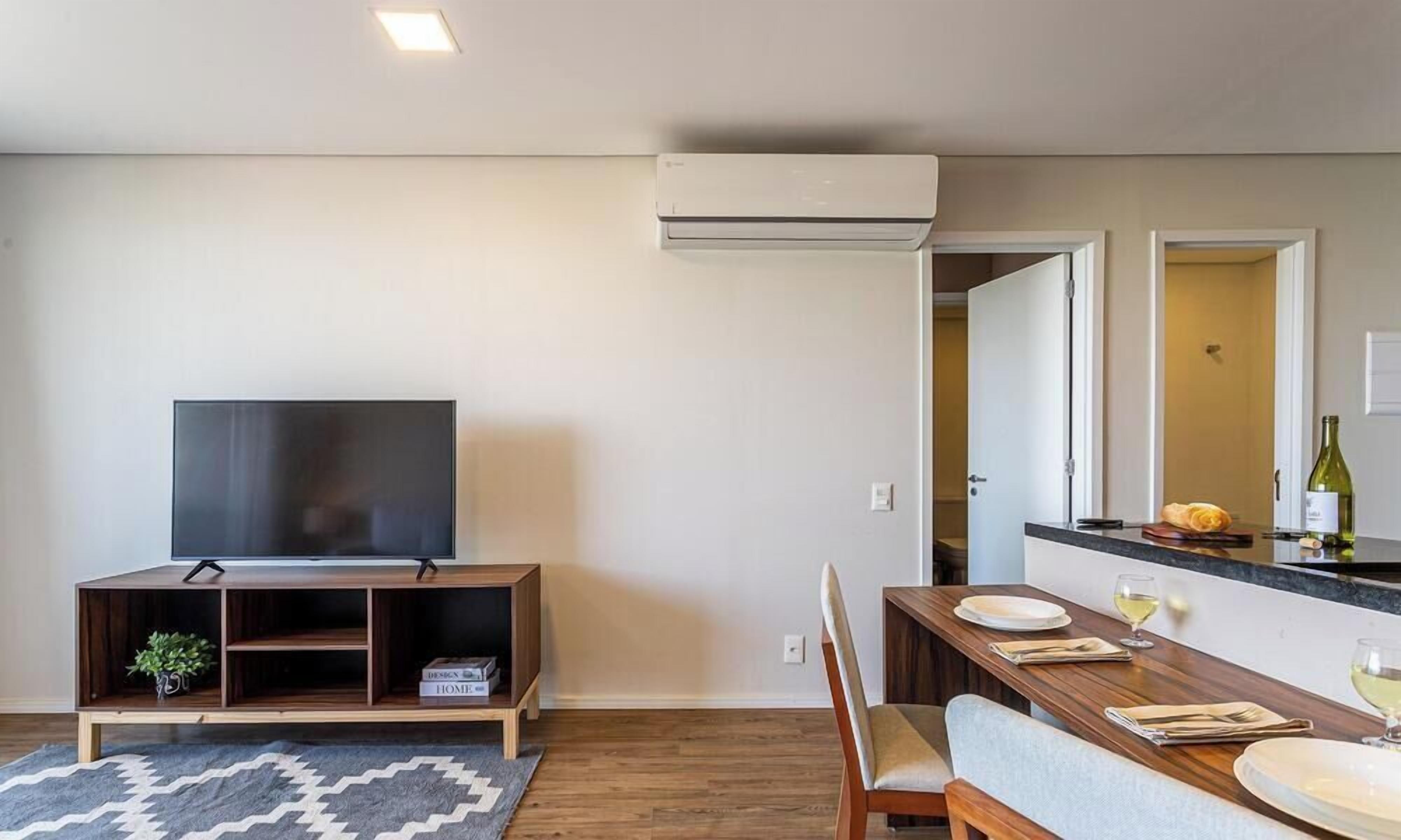 Una sala moderna con televisor de pantalla plana, mueble de madera para entretenimiento, mesa de comedor con sillas y una zona de cocina con mostrador y botella de vino.