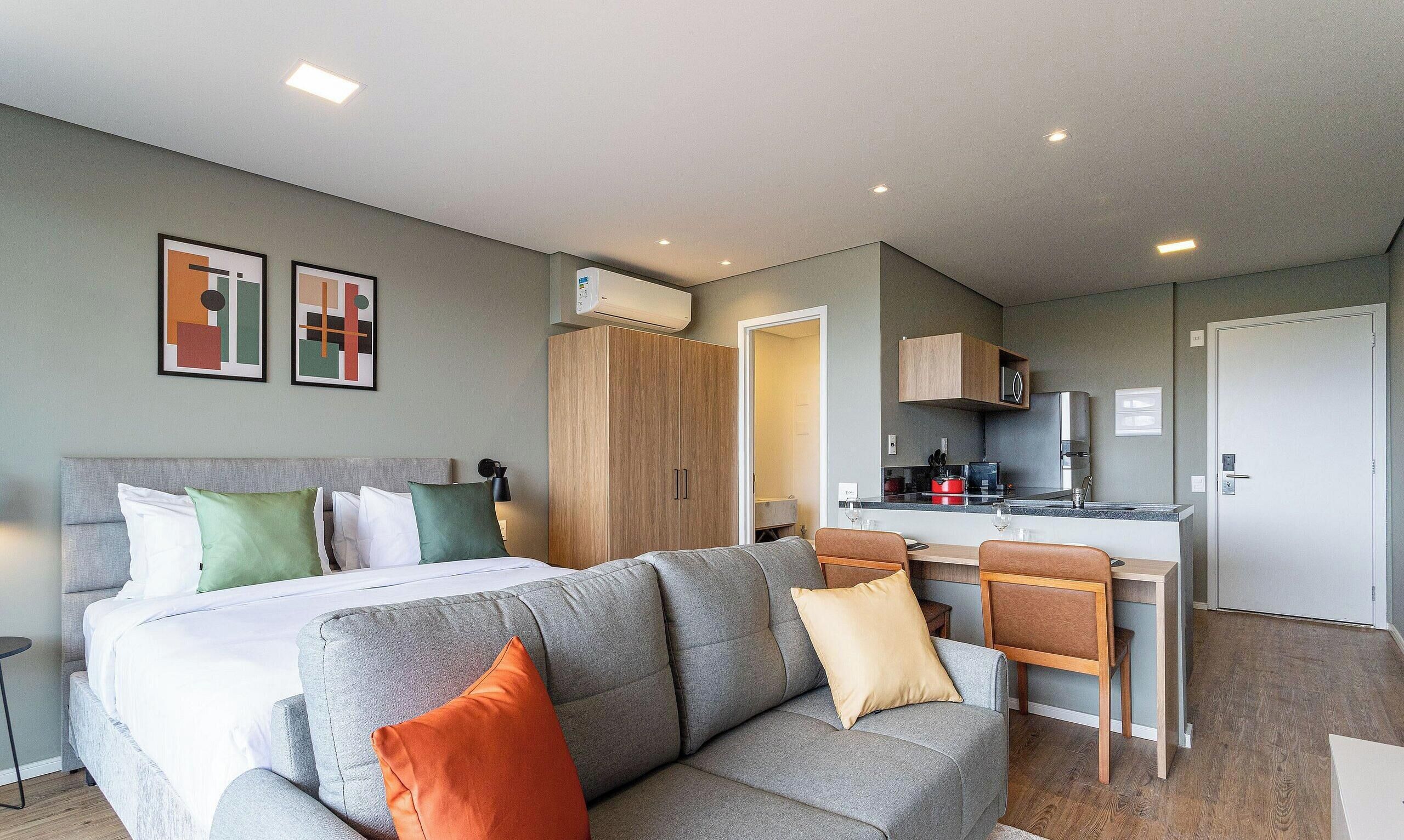 A modern hotel room with a bed, a grey sofa, a dining area, and a kitchen.