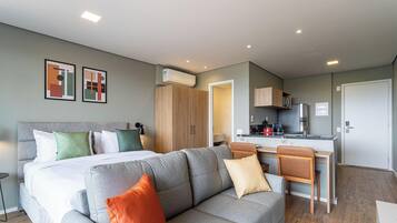 A modern hotel room with a bed, a grey sofa, a dining area, and a kitchen.