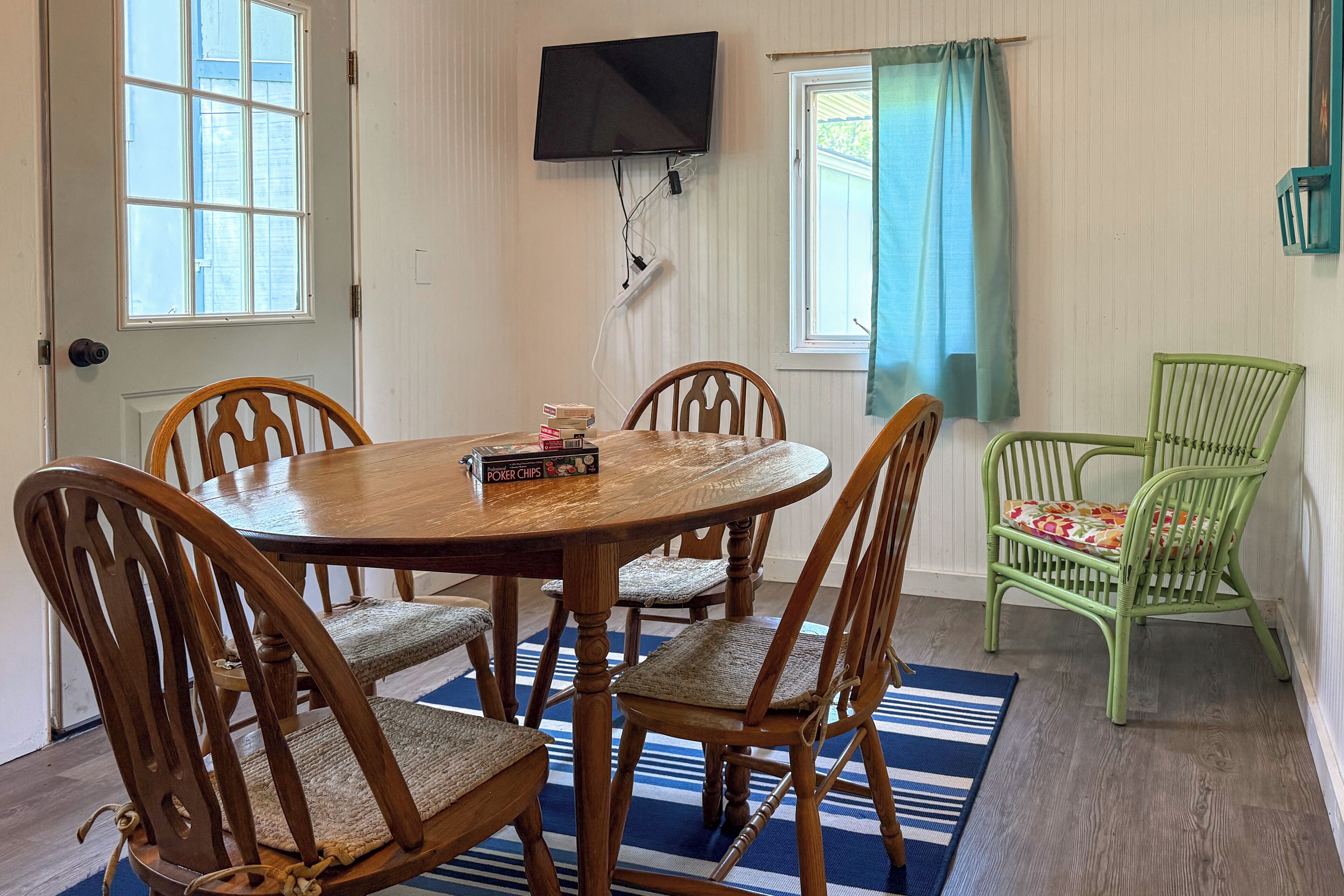 A dining area with a round wooden table, four chairs, a television mounted on the wall, and a window with curtains.