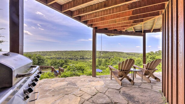 Een overdekt terras met een stenen patio, een barbecue en Adirondack-stoelen met uitzicht op een bebost landschap.