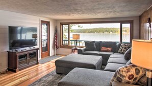 Una sala de estar con una televisión grande, un sofá, otomanas y vistas a un lago.