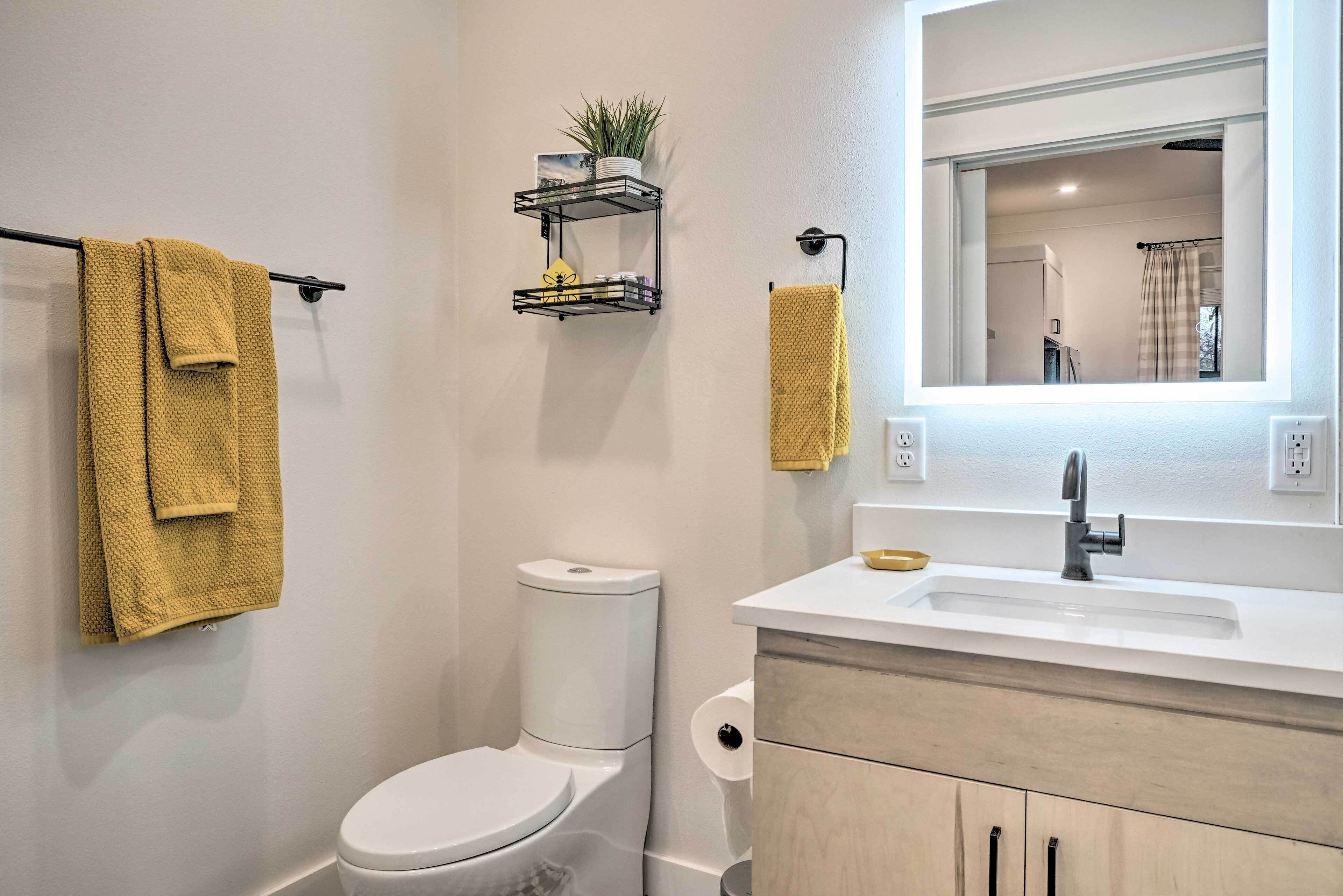 Un baño moderno con inodoro, lavamanos y espejo. Hay toallas colgadas en un perchero y una repisa con una planta.