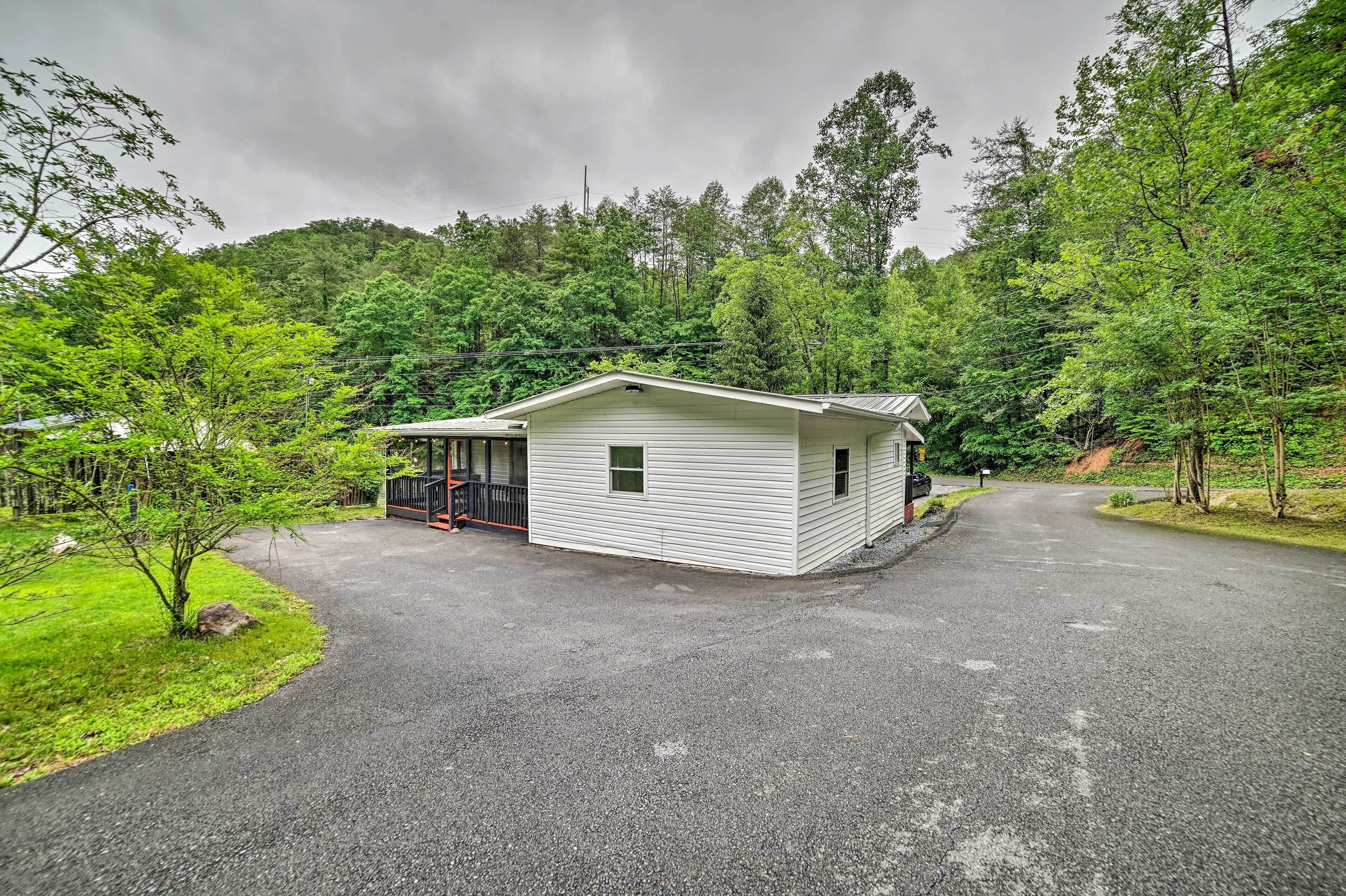 A small, single-story cabin with a porch, surrounded by lush greenery and a forested hillside.