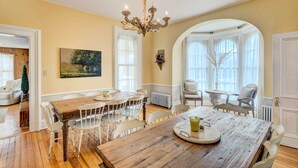 A dining area with a wooden table and white chairs, a chandelier, and large windows with white curtains.