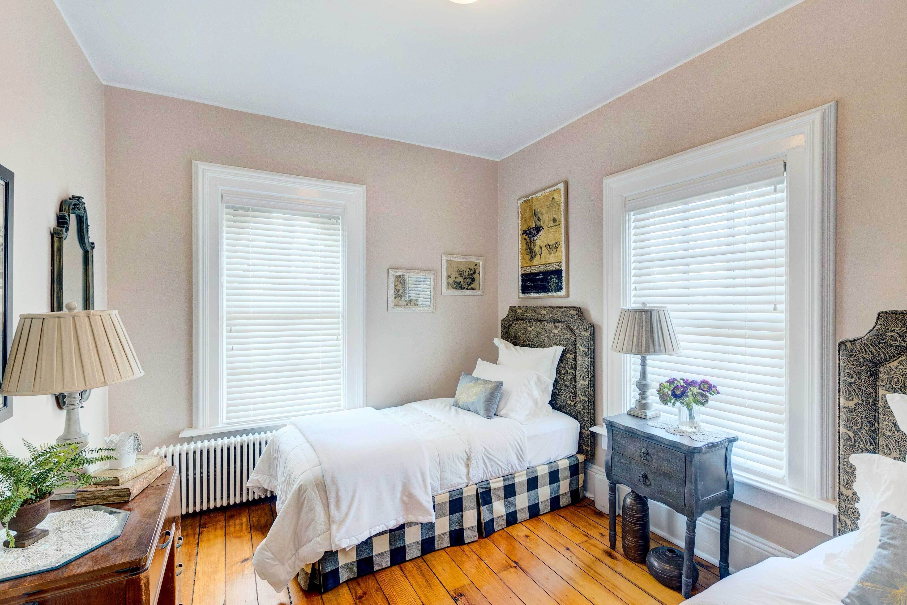 A bedroom with two beds, a radiator, a wooden dresser, and a nightstand with a lamp and flowers.