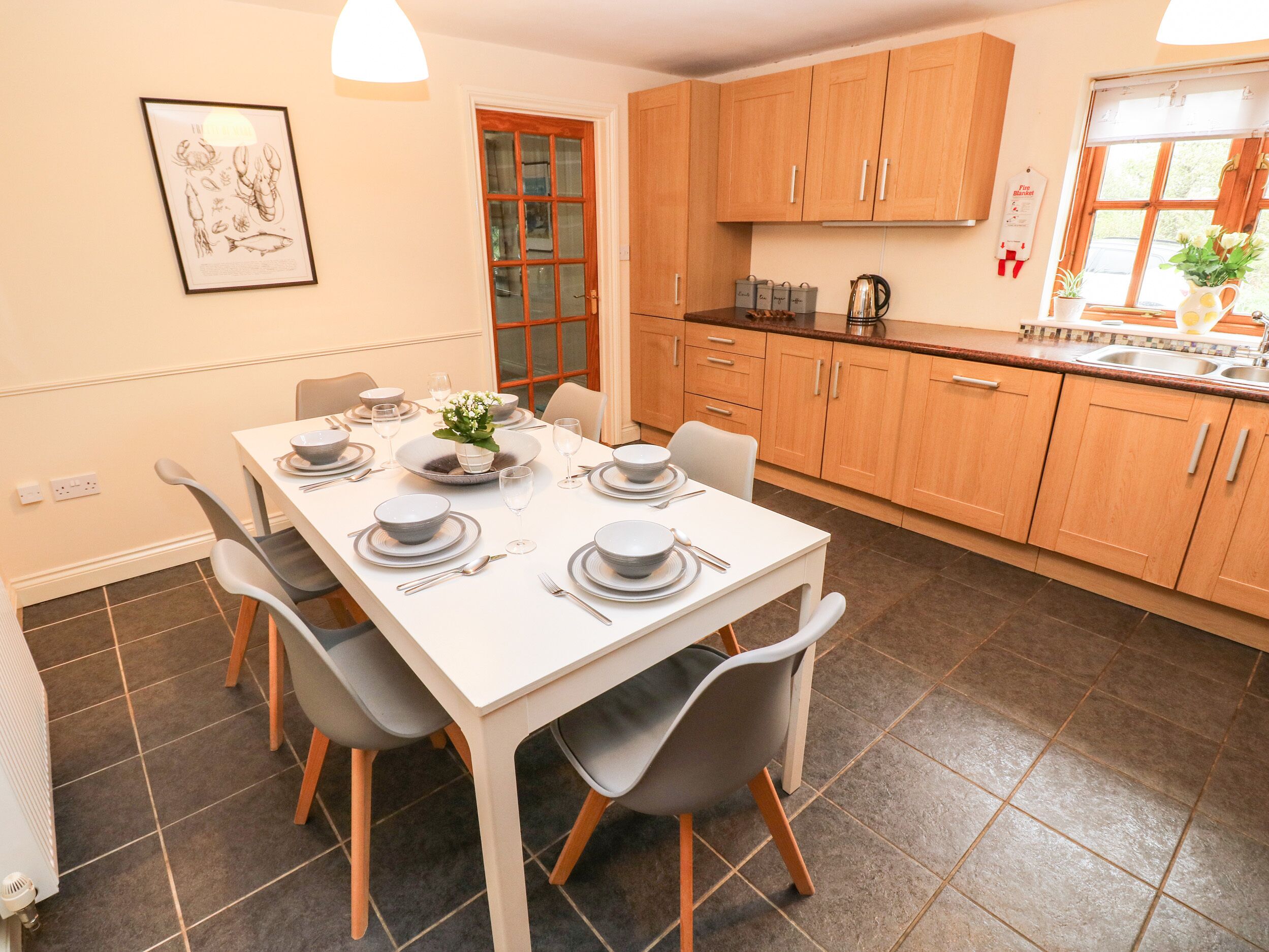 A dining area with a white table set for four, grey chairs, wooden cabinets, and a kitchen area with a window.