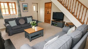 A living room with a grey sofa, a coffee table with a vase, a television on a stand, and a staircase. - Ty Rhosyn (Haverfordwest)