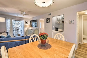 A dining area with a wooden table, chairs, and a vase of red flowers. There is a ceiling fan, a television mounted on the wall, and a kitchen area visible through an open doorway.