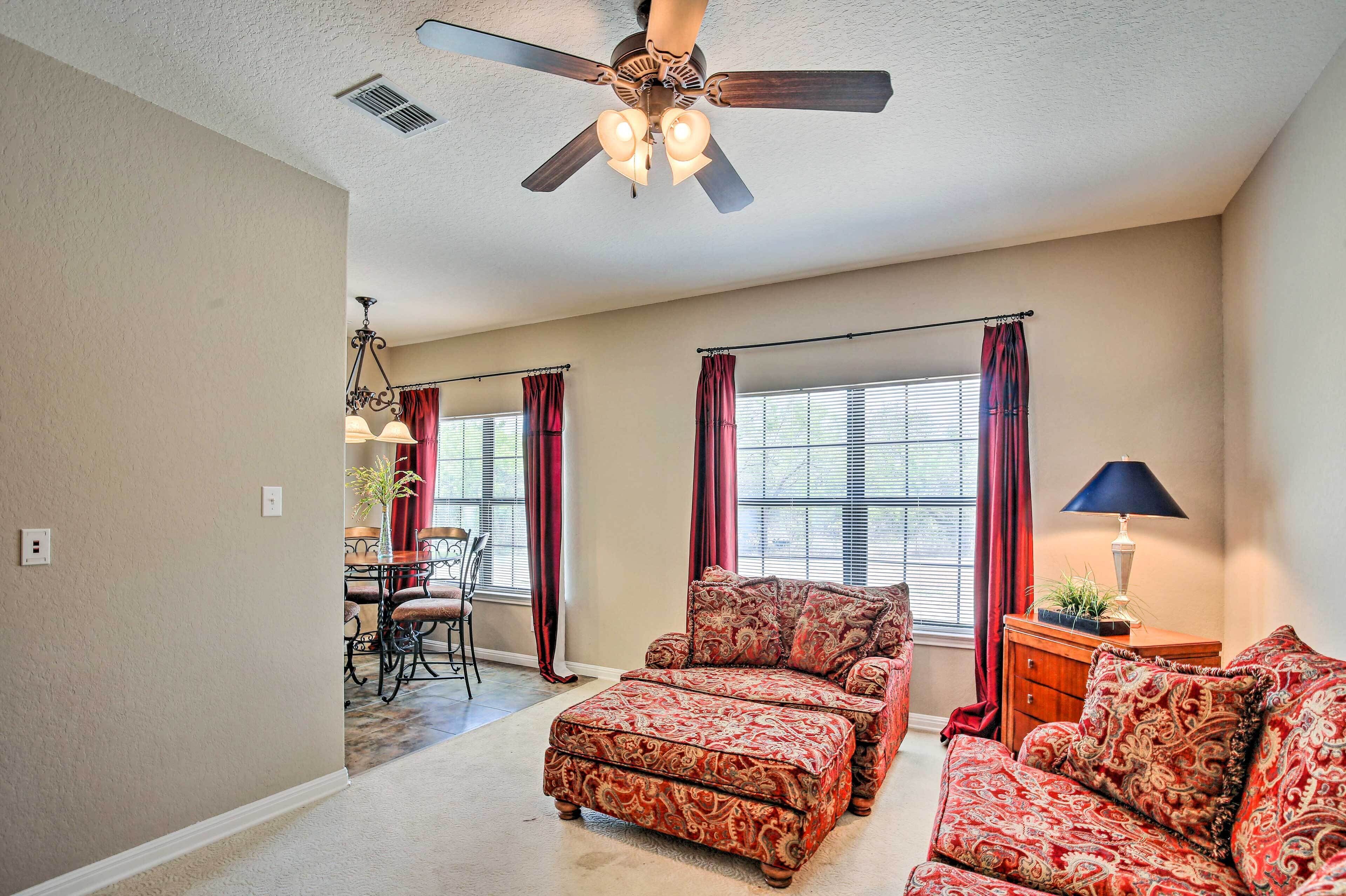 A spacious living room with a ceiling fan, a dining area with a chandelier, and a view through large windows.