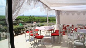 Un área de comedor al aire libre con carpas, mesas y sillas blancas, cojines rojos y vistas a un paisaje verde.