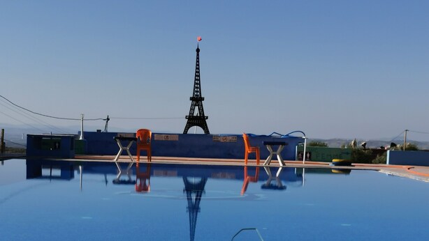 Una piscina con el reflejo de la Torre Eiffel, rodeada de sillas naranjas y una valla azul.