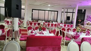 Un salón de banquetes con filas de mesas y sillas, ventanas amplias y una zona de escenario.