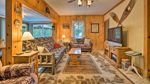 Un salón acogedor de estilo cabaña con paredes de madera, un ventilador de techo, un televisor y un ventanal grande.
