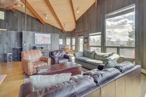 Ein geräumiges Wohnzimmer mit hohen Decken, Holzbalken, großen Fenstern und verschiedenen Sitzgelegenheiten, darunter eine Couch, Sessel und ein gemustertes Sessel.