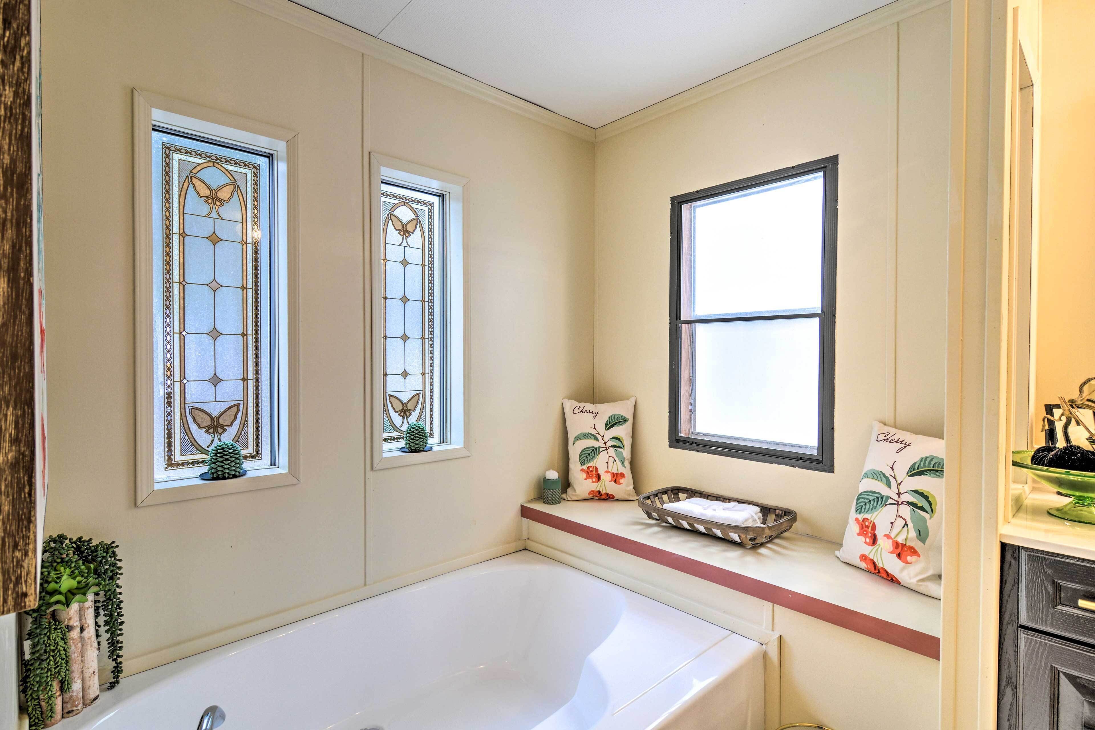 A bathroom with a bathtub, two windows with decorative glass, a shelf with decorative pillows, and a small window with a plant.