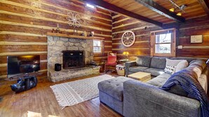 A cozy log cabin interior with a stone fireplace, a television, a comfortable sofa, and a rustic wooden ceiling. - Cozy Log Cabin: 6 Mi to Great Smoky Mtns NP! (Townsend)