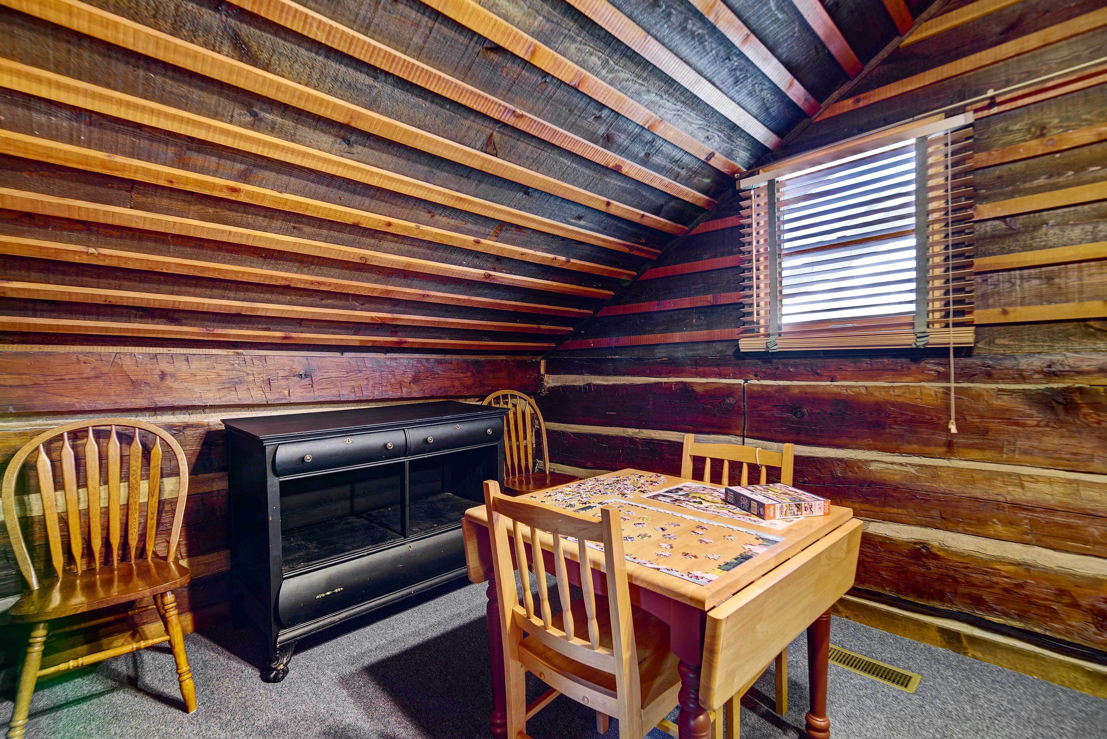 A cozy cabin room with a wooden ceiling, a fireplace, a dining table with chairs, and a window with blinds.