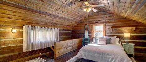 Ein gemütliches Schlafzimmer in einer Blockhütte mit Holzboden, einem Bett mit weißer Bettwäsche, einem Nachttisch und einem Fenster mit Vorhängen.