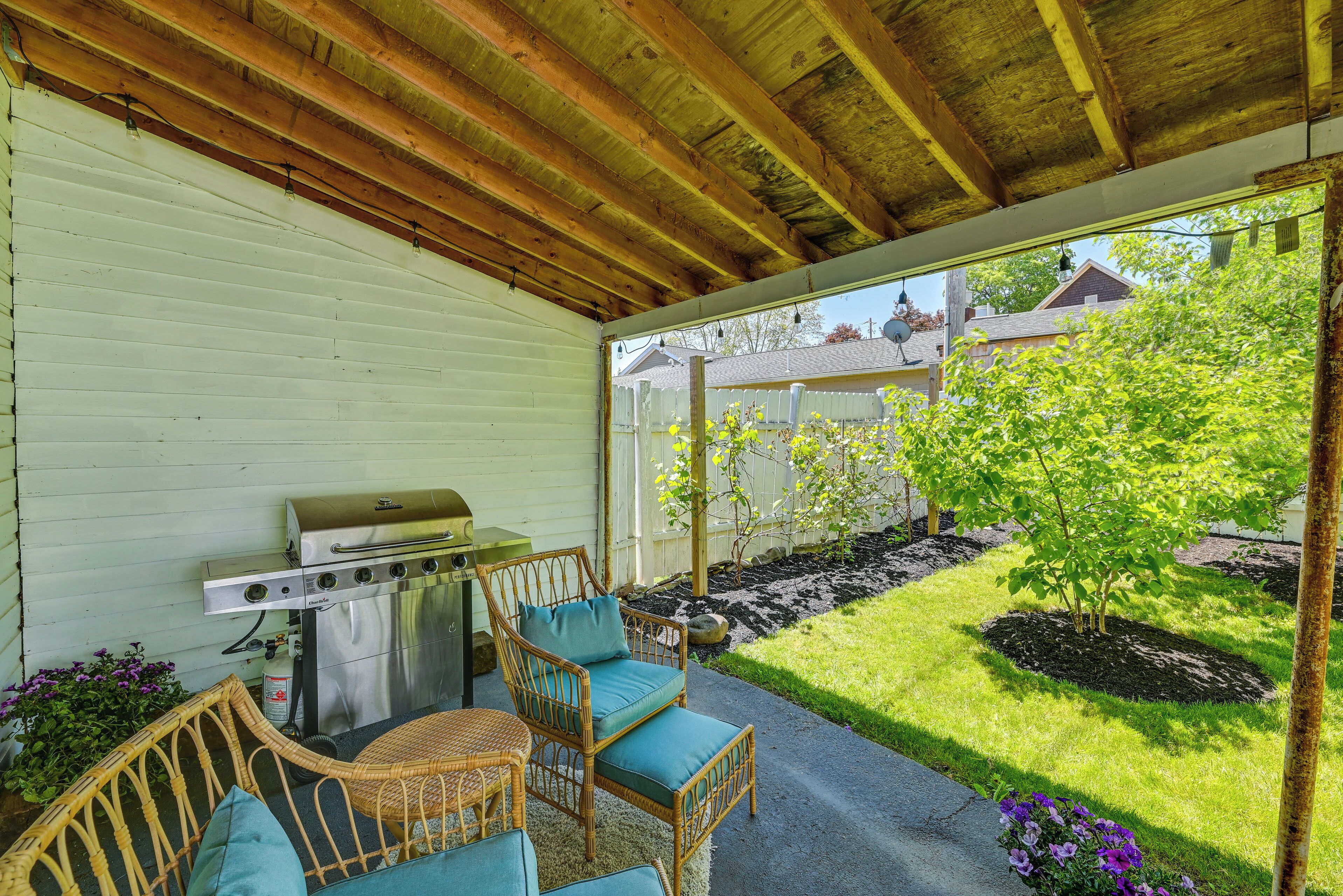 Une terrasse couverte équipée d’un barbecue, de meubles en osier et offrant une vue sur un jardin avec une pelouse et des arbres.