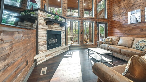 A log cabin interior with a stone fireplace, leather sofa, and large windows. - Sleek Cabin w/ Hot Tub: 11 Mi to Broken Bow Lake! (Broken Bow)