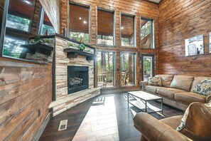 A log cabin interior with a stone fireplace, leather sofa, and large windows. - Sleek Cabin w/ Hot Tub: 11 Mi to Broken Bow Lake! (Broken Bow)