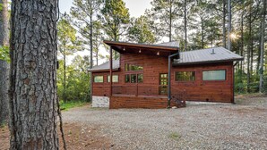 A rustic cabin with a wooden exterior, large windows, and a gravel driveway. - Sleek Cabin w/ Hot Tub: 11 Mi to Broken Bow Lake! (Broken Bow)