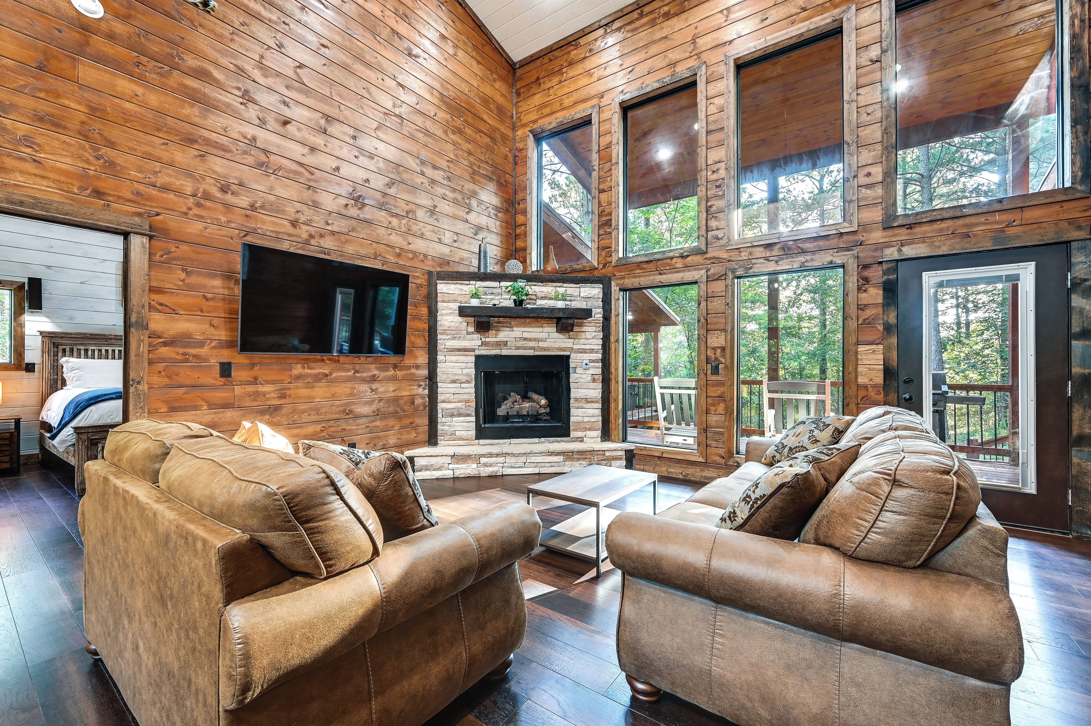 A log cabin interior with a fireplace, leather sofas, and large windows offering a view of the forest.