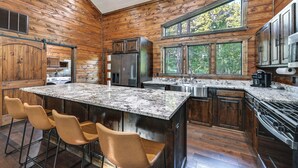 A modern kitchen with wooden walls, a large island with bar stools, stainless steel appliances, and a view of the forest through large windows. - Sleek Cabin w/ Hot Tub: 11 Mi to Broken Bow Lake! (Broken Bow)