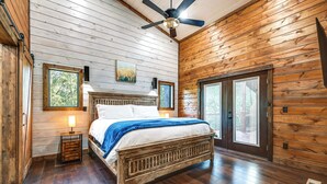 A wooden-paneled bedroom with a bed, a ceiling fan, and a large window. - Sleek Cabin w/ Hot Tub: 11 Mi to Broken Bow Lake! (Broken Bow)