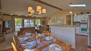 Un coin repas avec une table en bois dressée pour six personnes, une cuisine avec des meubles blancs et une grande fenêtre donnant sur l’extérieur.
