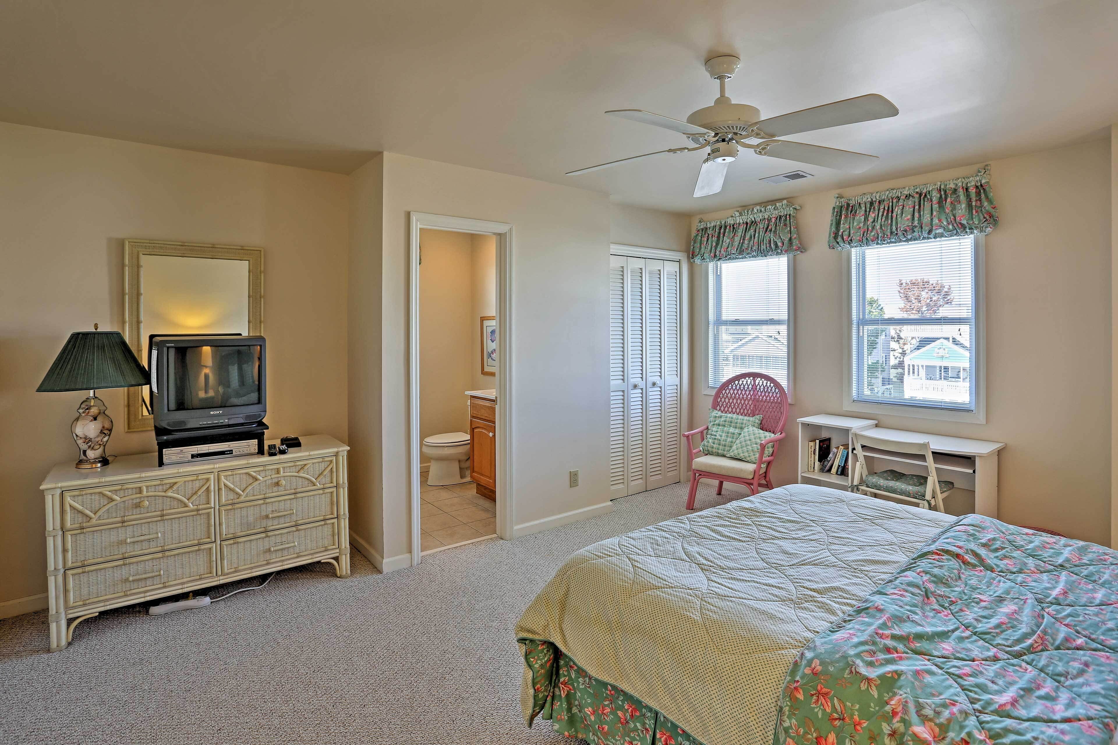 A bedroom with a bed, a television on a dresser, a ceiling fan, and a window with shutters.