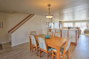 A dining area with a wooden table and chairs, a kitchen in the background, and a staircase.