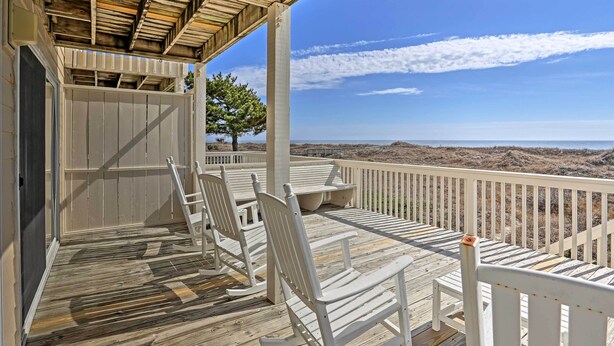 A wooden deck with white chairs and a table, overlooking a beach.