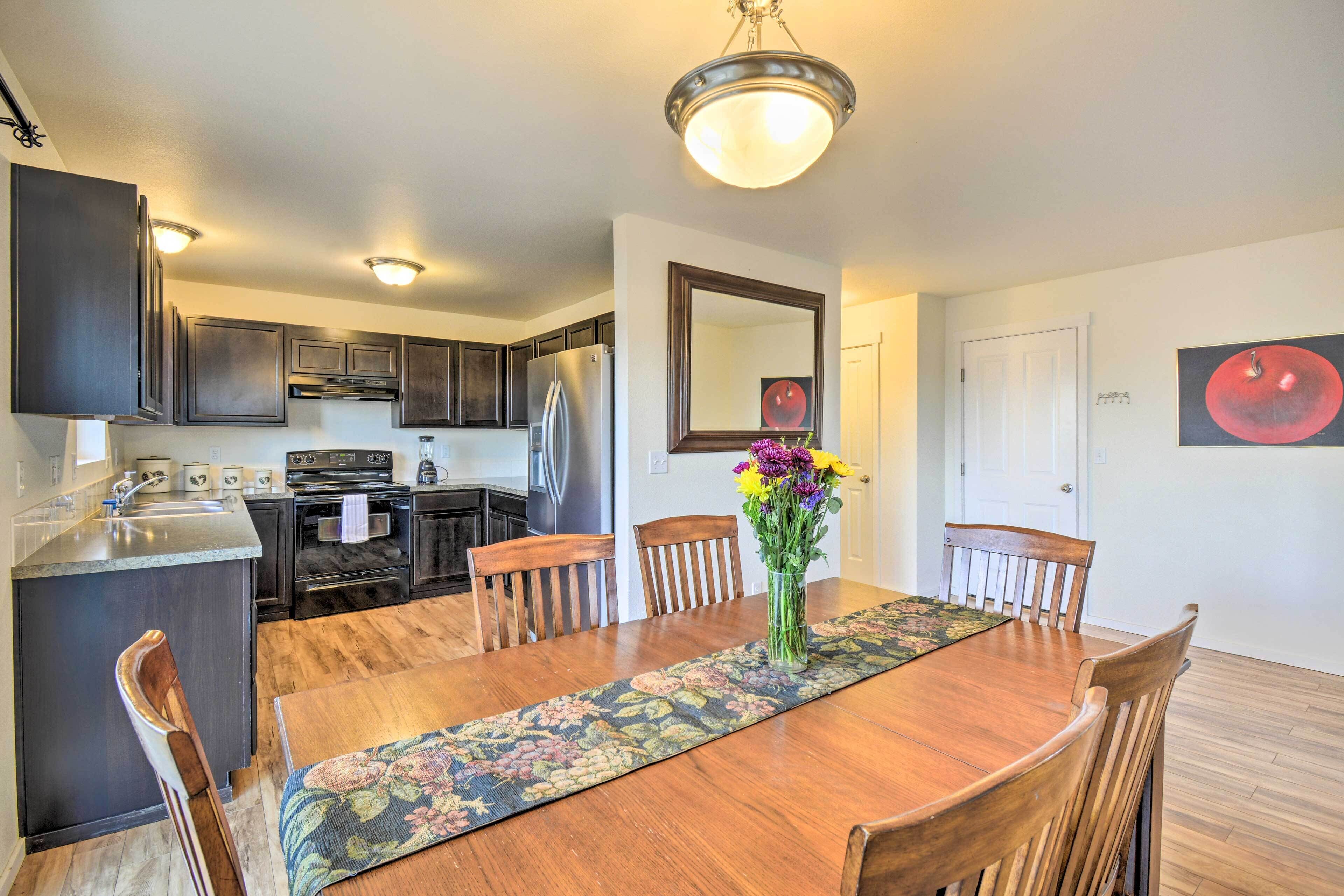 A dining area with a wooden table, chairs, a vase with flowers, and a kitchen in the background.