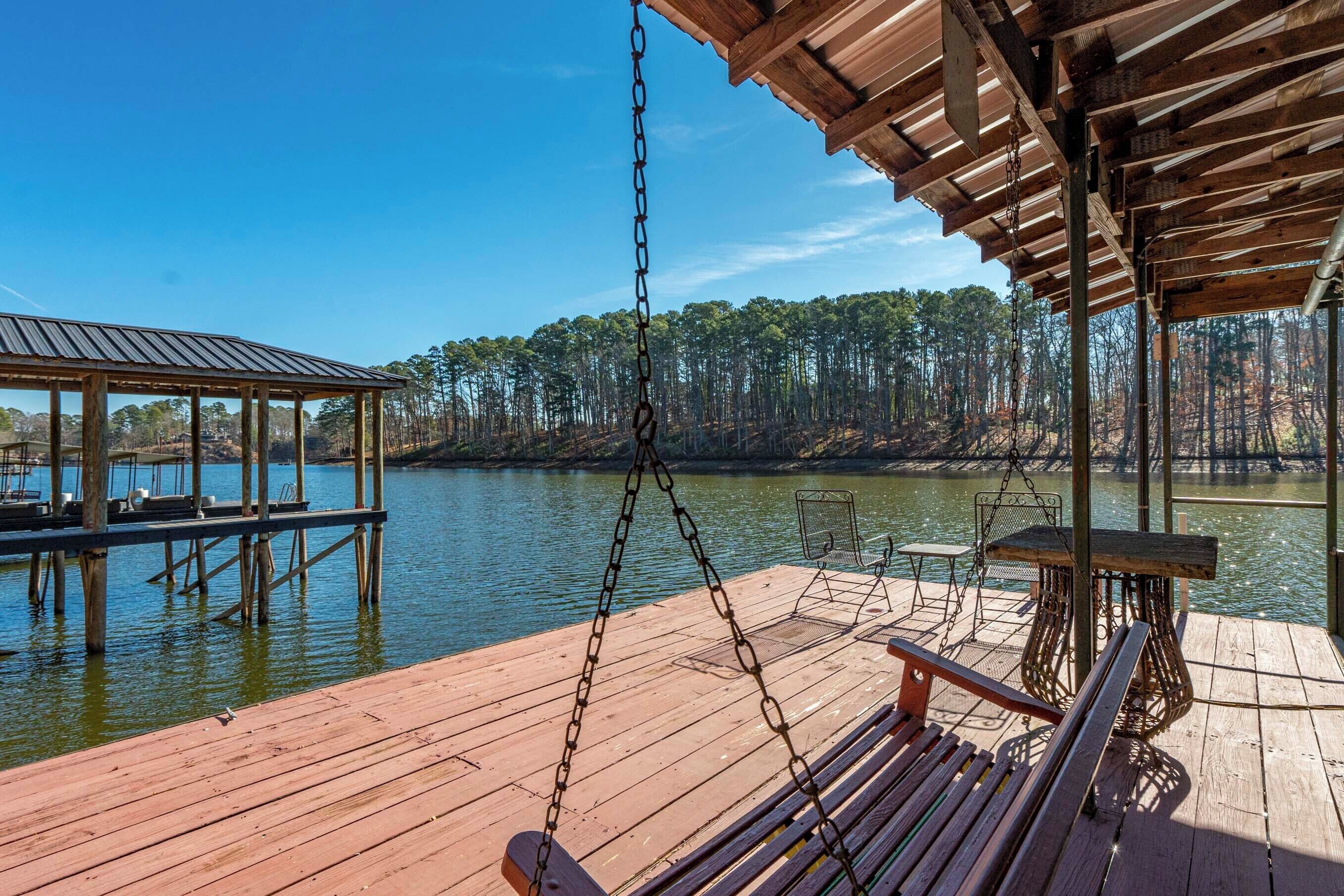 A wooden dock with a swing chair and a table, overlooking a calm lake with trees in the background.