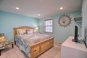 A bedroom with a wooden bed, a nightstand with a lamp, a window with blinds, a wall-mounted mirror, and a television on a stand.