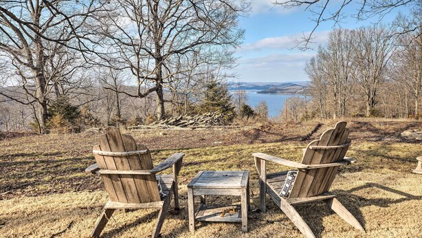 Twee Adirondack stoelen en een klein houten tafeltje staan op een gazon met uitzicht op een meer en bomen.