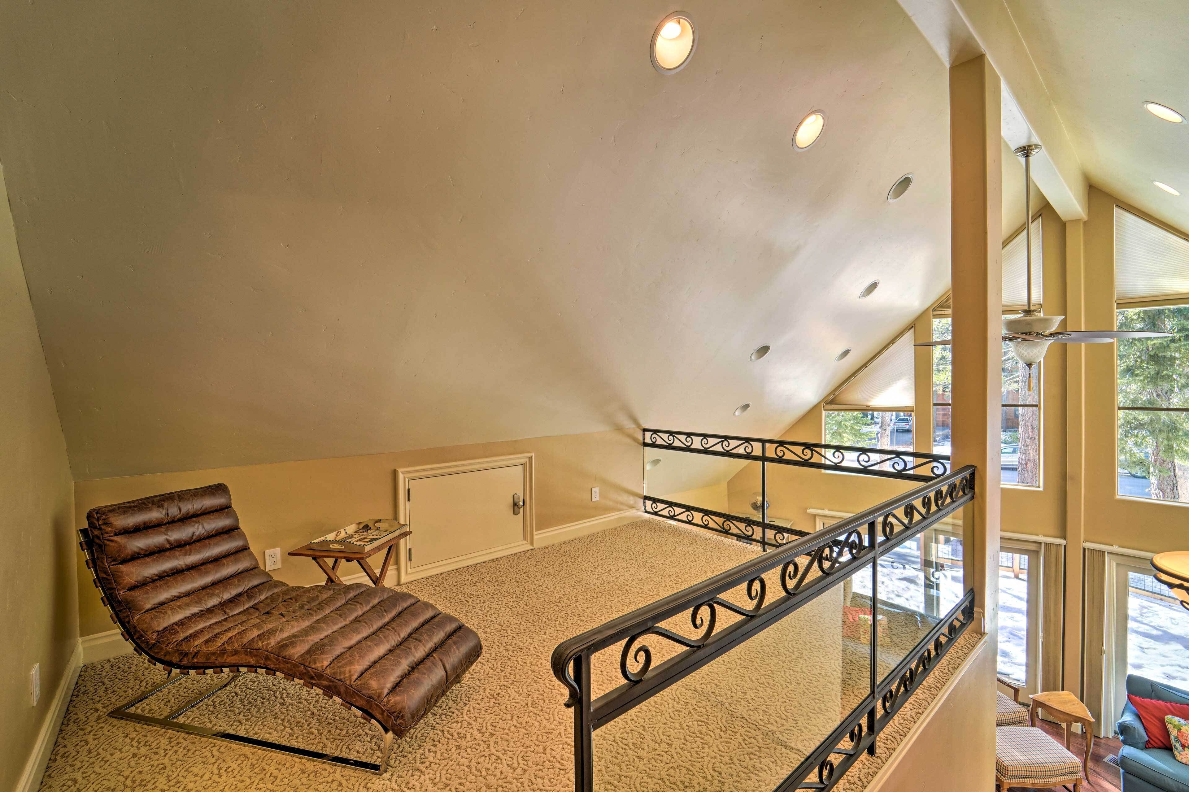 A spacious loft bedroom with a large window, a chaise lounge, and a staircase with a railing.