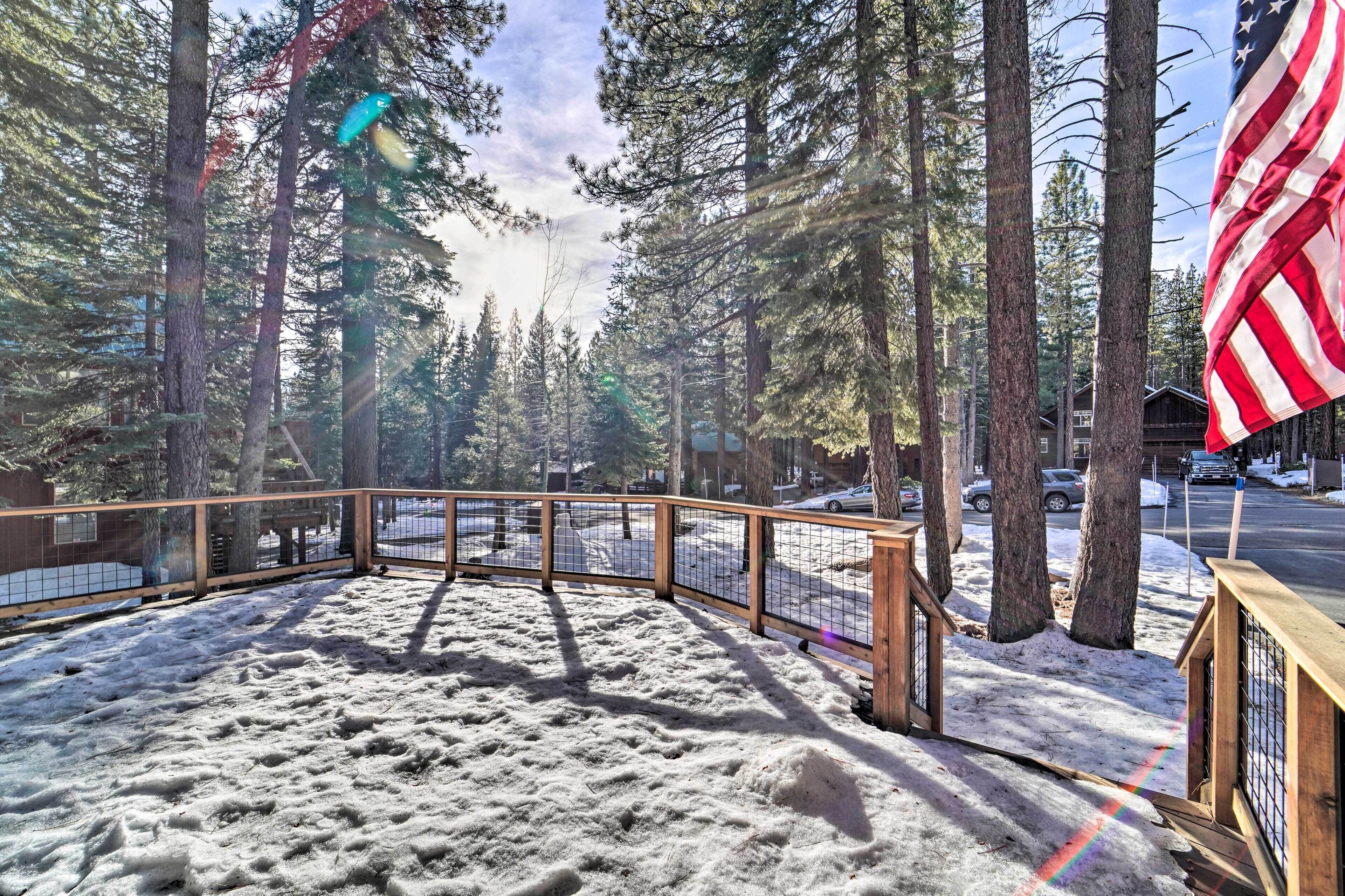Een sneeuwbedekt terras met houten reling, omringd door hoge dennen en een Amerikaanse vlag.