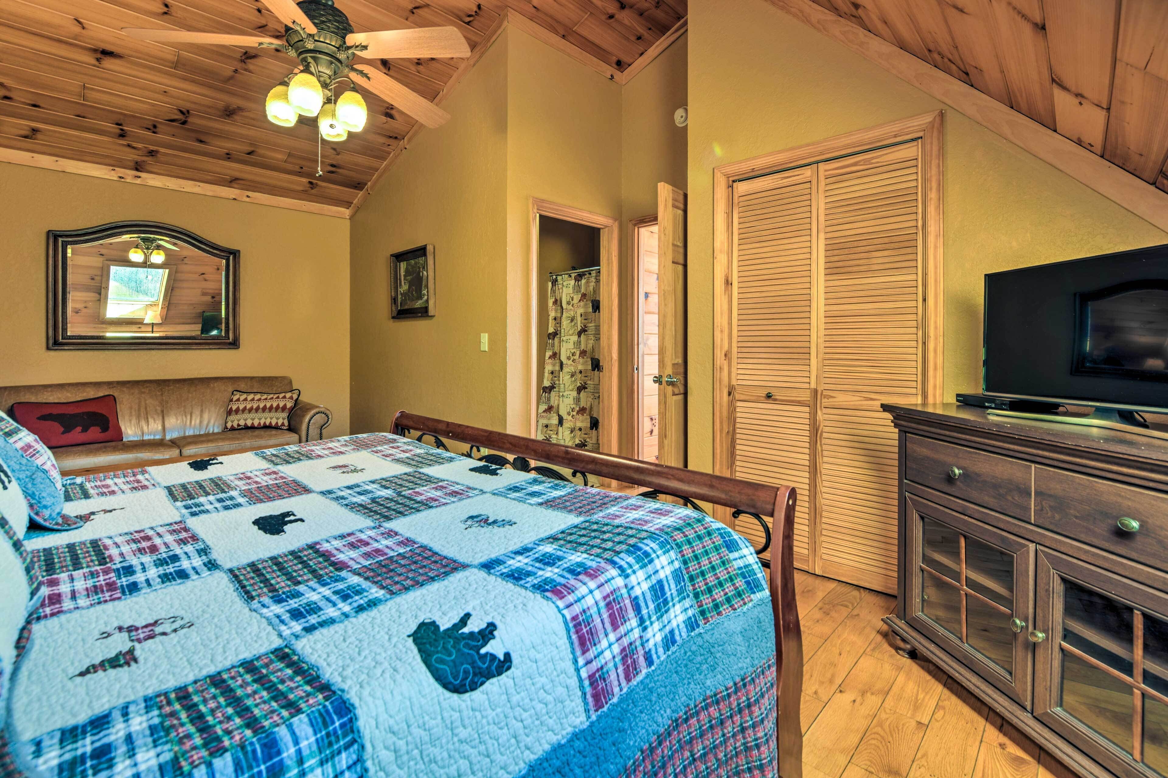 Un dormitorio con una cama grande, techo de madera, televisor sobre un mueble y una puerta que da a otra habitación.