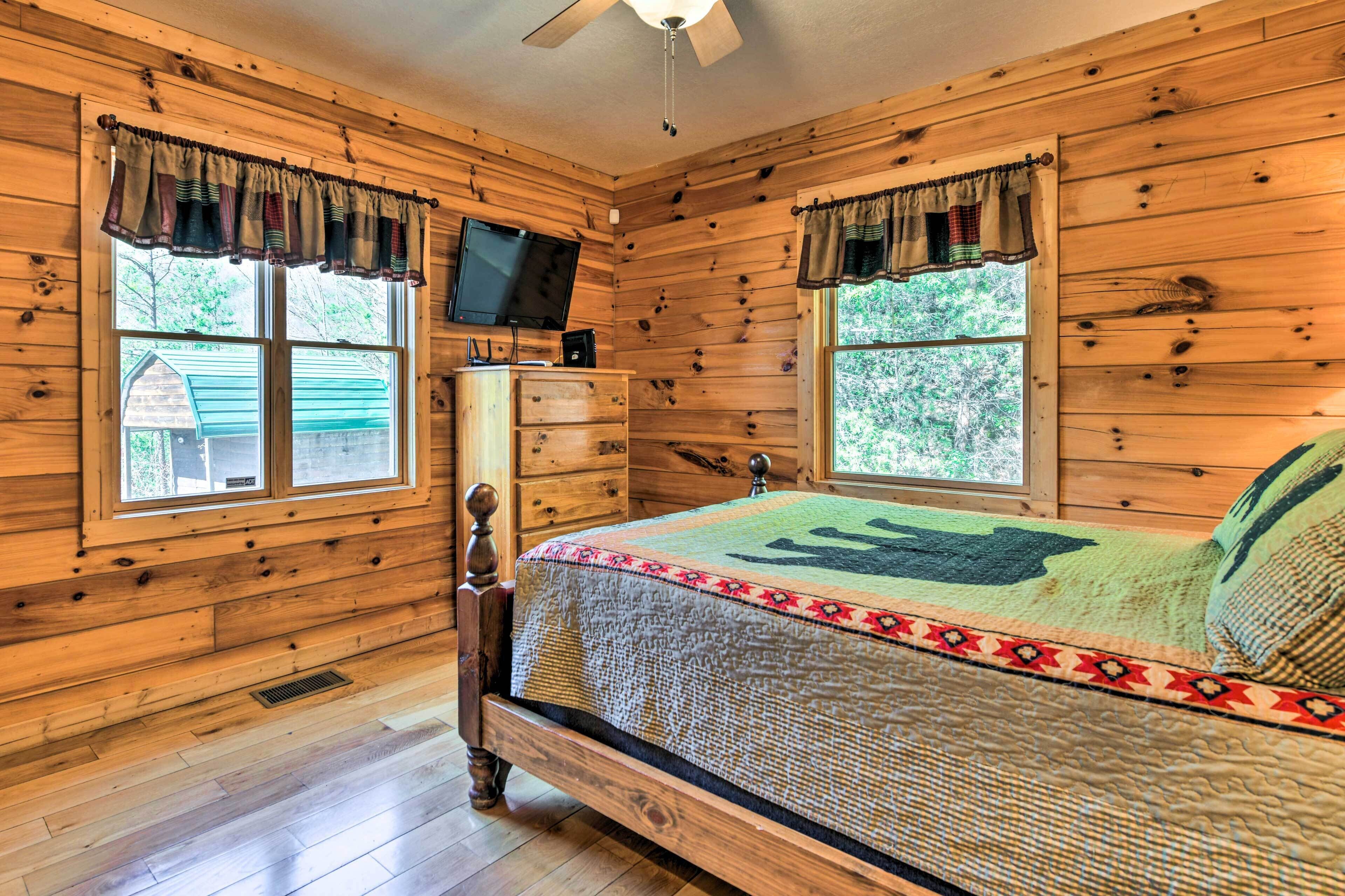 Un dormitorio de cabaña de madera con cama, cómoda, televisor y ventanas.