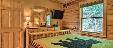 A wooden cabin bedroom with a bed, a television, a window with a view of trees, and a door.
