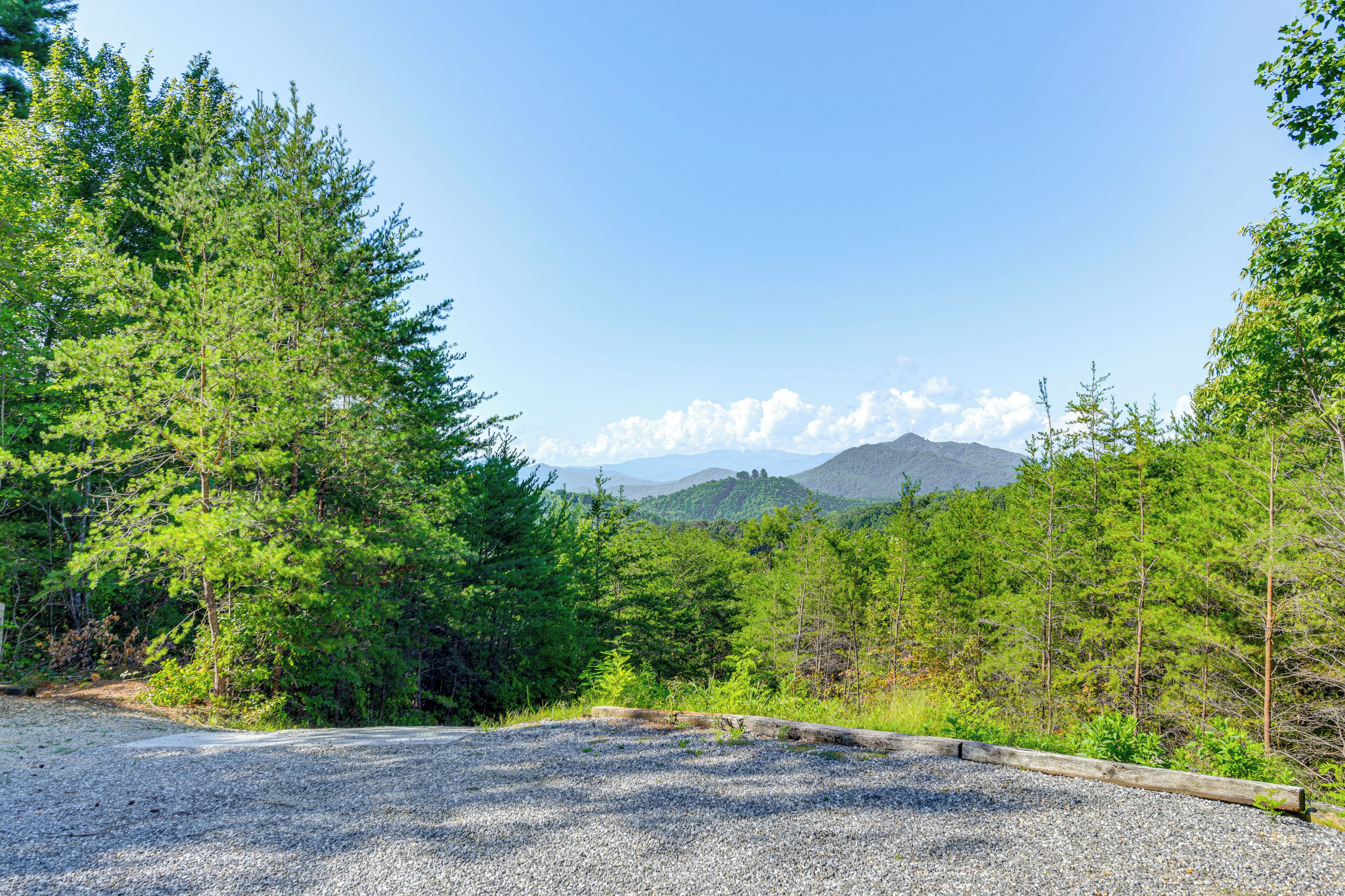 A gravel parking area with a wooden fence, surrounded by dense green trees and a mountain in the background.