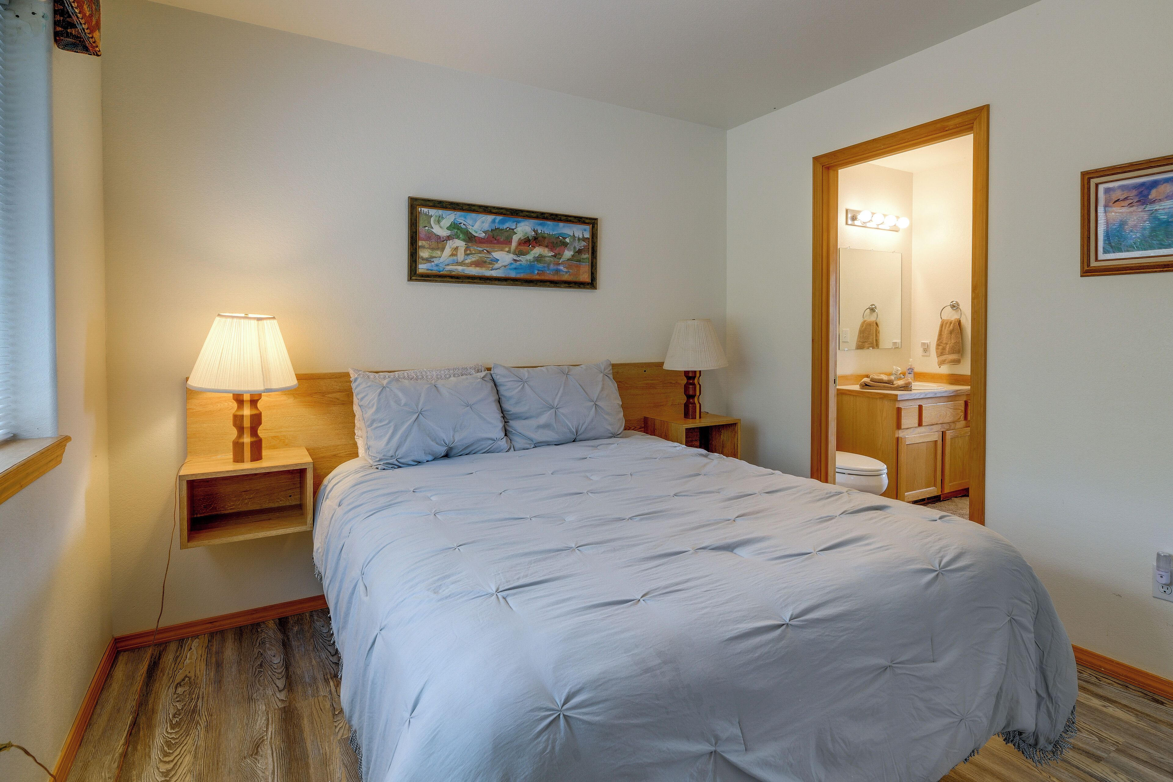 A neatly arranged bedroom with a bed, two bedside lamps, a nightstand, a wooden headboard, a painting, and a bathroom visible through an open door.