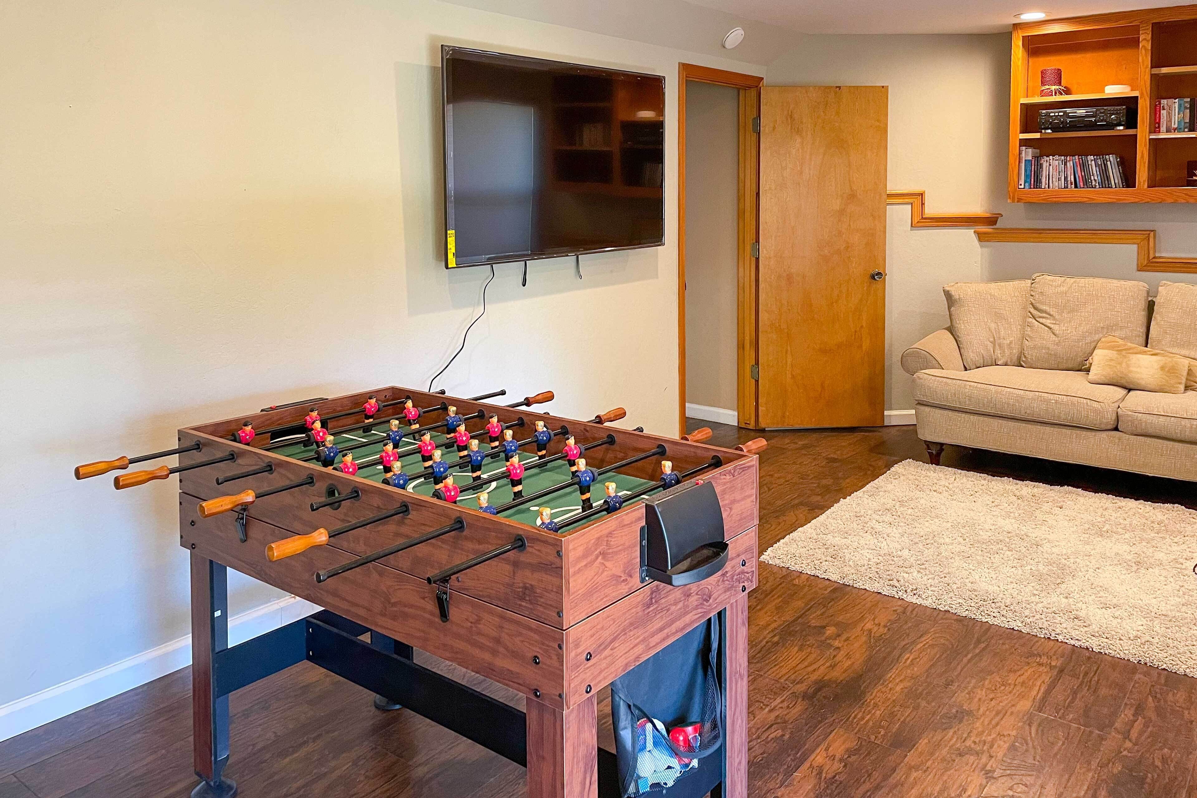 A room with a foosball table, a television mounted on the wall, a sofa, and a bookshelf.