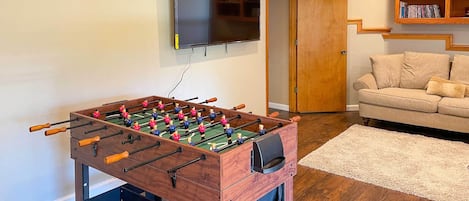A room with a foosball table, a television mounted on the wall, a sofa, and a bookshelf.