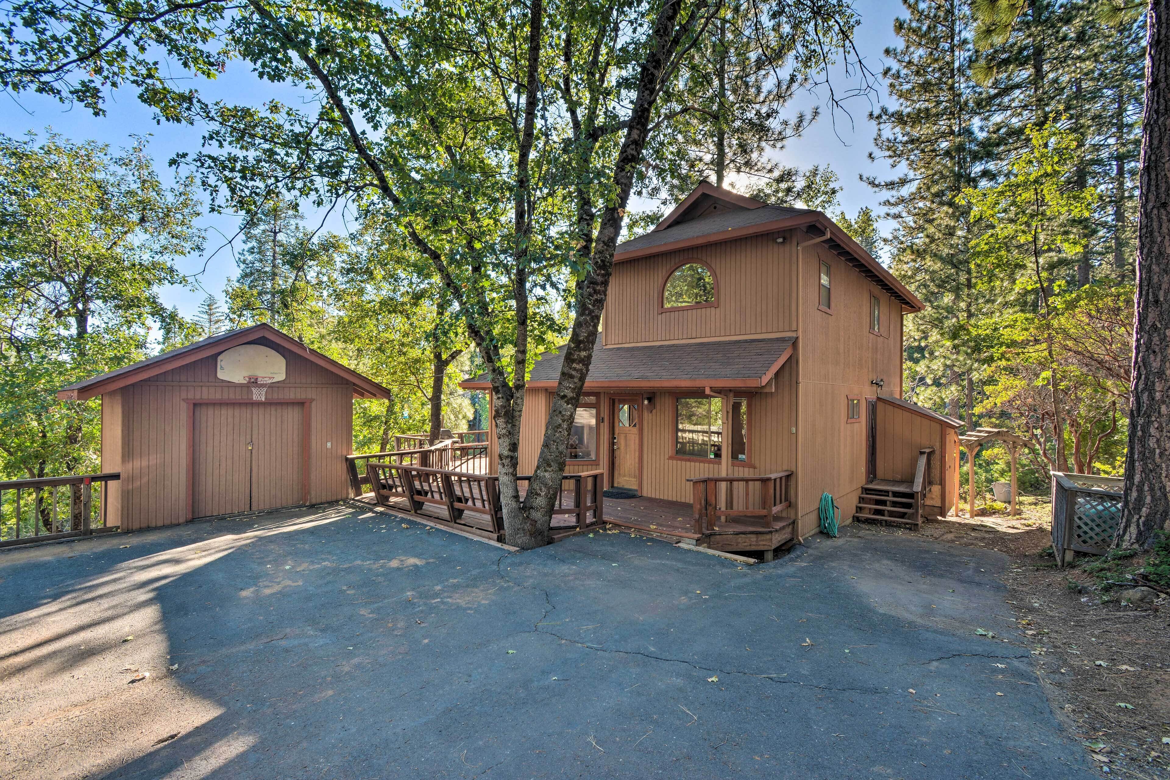 Une cabane en bois à deux étages, dotée d’un porche couvert et d’un garage, entourée d’arbres.