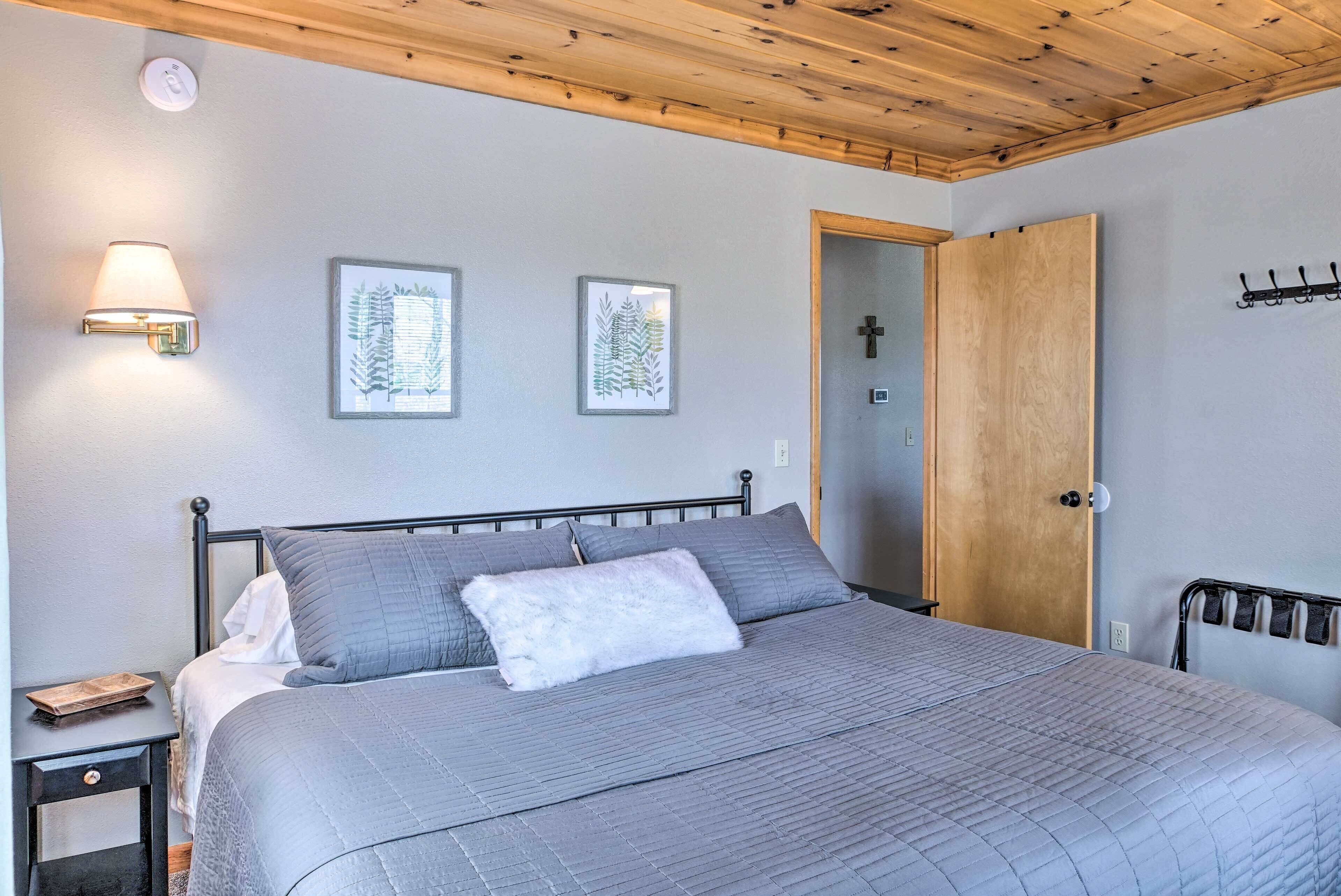 A bedroom with a bed, a nightstand, a wall-mounted lamp, framed botanical prints, a wooden door, and a coat rack.